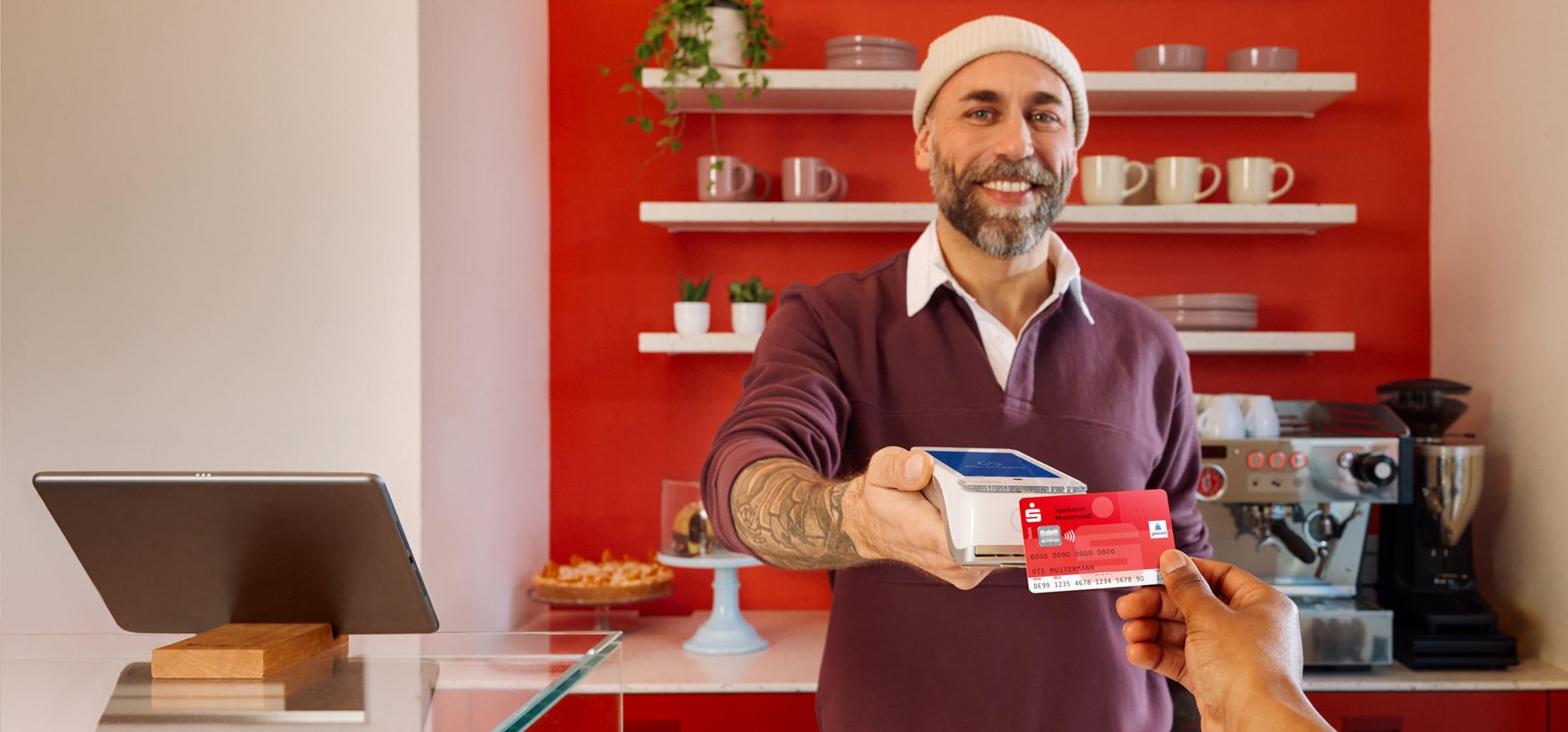 Lächelnder Barista mit Mütze reicht ein Kartenlesegerät über den Tresen, während eine Kundin mit einer roten Sparkassen-Girocard kontaktlos bezahlt