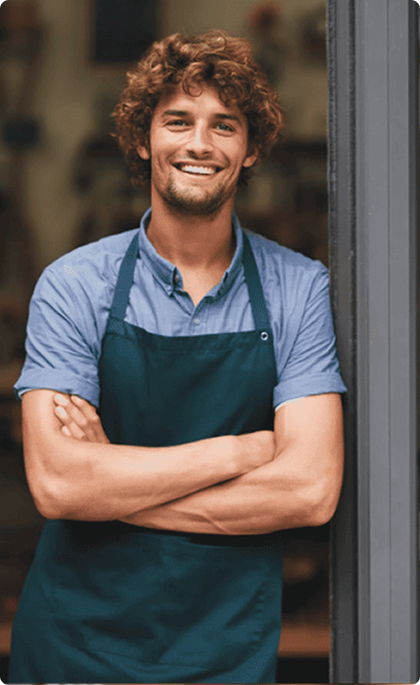 Junger Mann mit Schürze steht lächelnd in der Tür eines Geschäfts mit verschränkten Armen